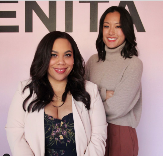 Two women entrepreneurs are photographed together. One is African American with long hair. She is sitting on a stool. The other woman is East Asian and she is standing next her with her arms crossed confidently.