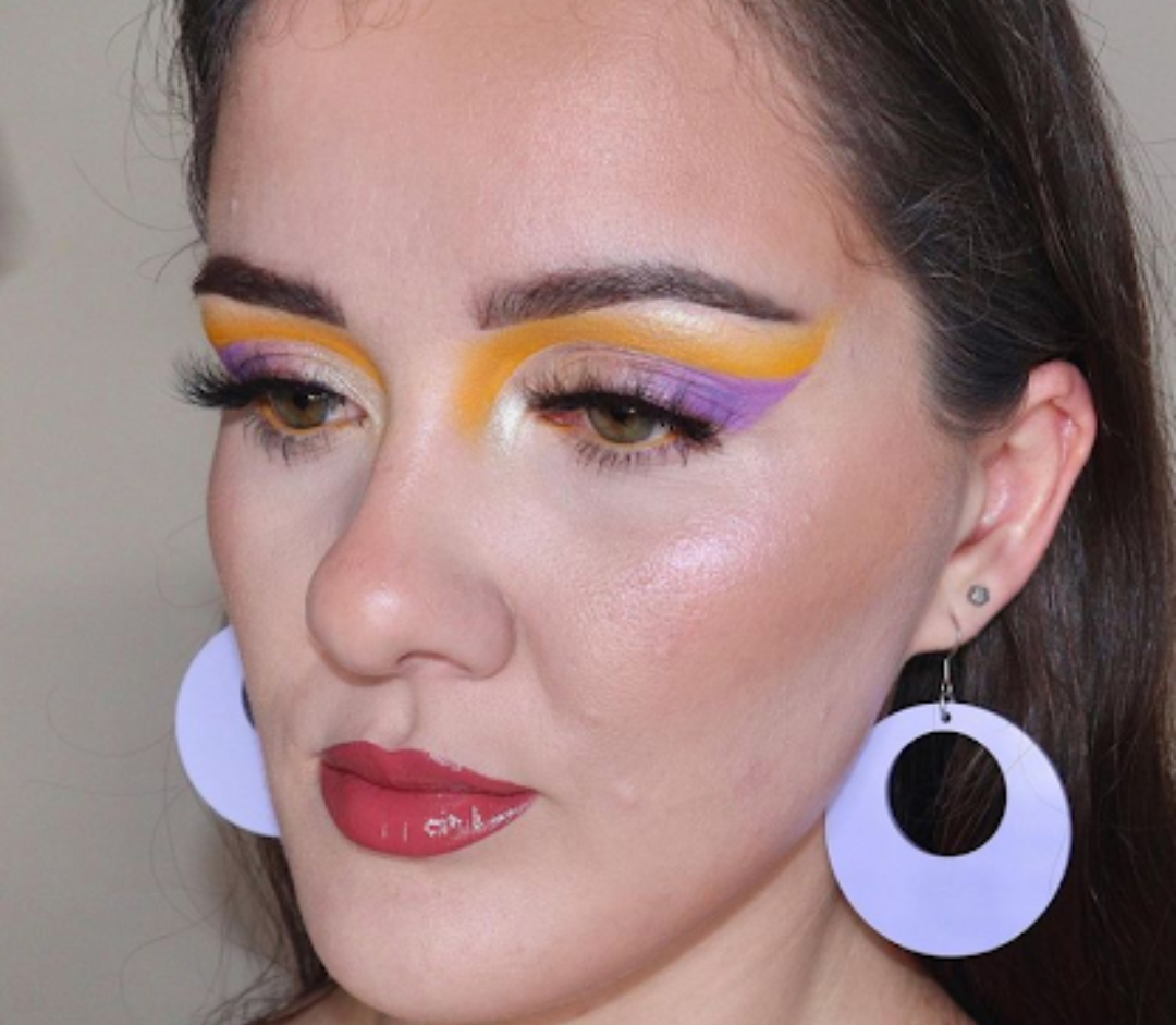 Chrissy is showing a makeup look that has lavender near her eyes in a wave form, and then the lavender is accentuated with a tangerine color. She is wearing circular earrings with a hole in the middle.